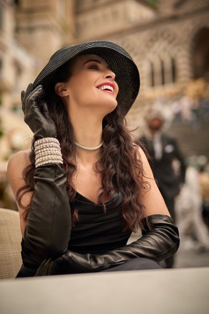 Elegant woman wearing black nappa leather opera gloves, styled with a wide-brimmed hat and pearl accessories, exuding luxury.