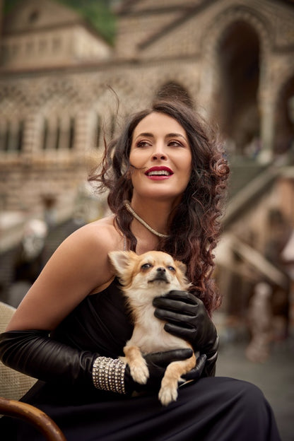 Elegant woman in black silk-lined nappa leather gloves, holding a small dog, exuding luxury in a scenic outdoor setting.