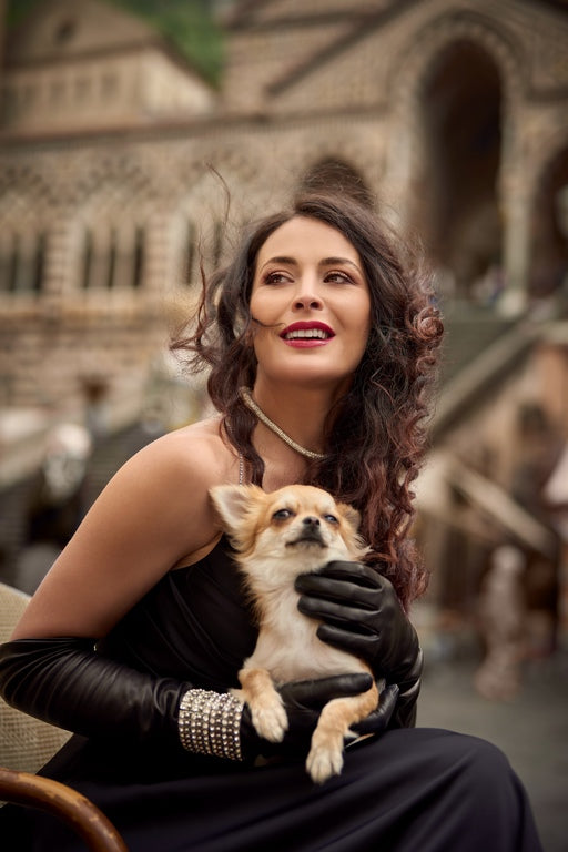Elegant woman in black silk-lined nappa leather gloves, holding a small dog, exuding luxury in a scenic outdoor setting.
