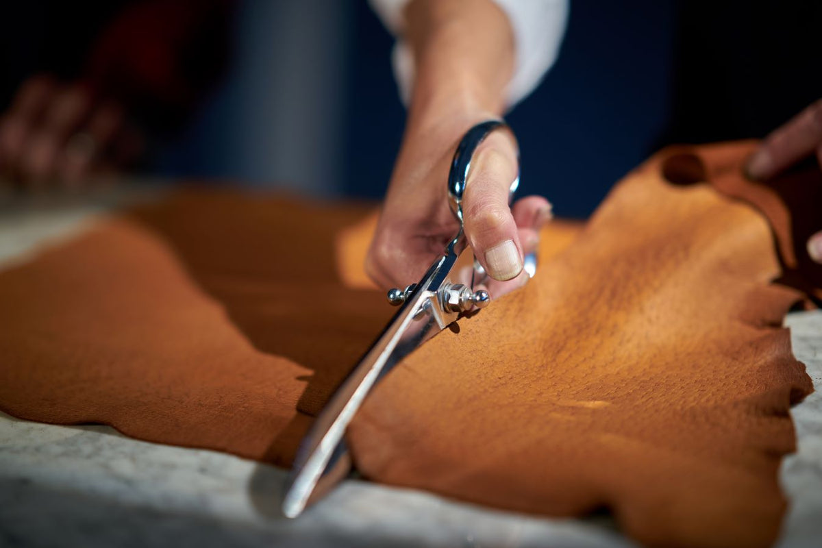 Artisan hands using scissors to cut rich brown leather, showcasing meticulous craftsmanship in glove-making.